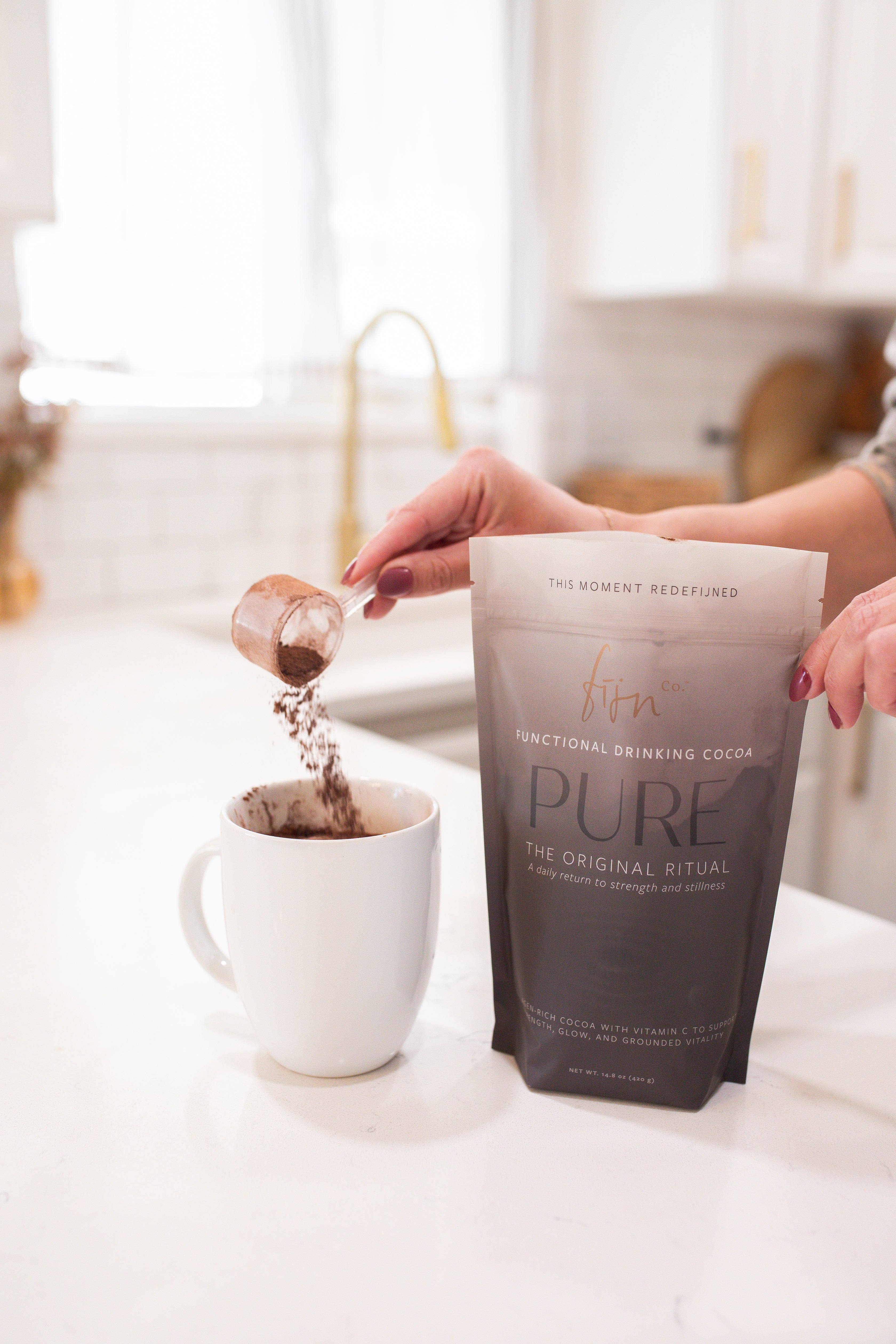 A hand scooping Fijn PURE cocoa collagen blend from an open bag, pouring it into a warm mug — your daily wellness ritual starts here.